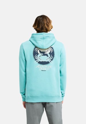 Person trägt einen hellblauen Hoodie mit einer Krabben-Grafik und dem Text "Adventure Division" auf dem Rücken, steht vor einem einfarbigen Hintergrund.