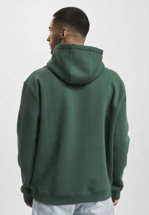 Mann mit kurzen Haaren, von hinten gesehen, trägt einen dunkelgrünen Kapuzenpullover und helle Hosen vor einem schlichten Hintergrund.