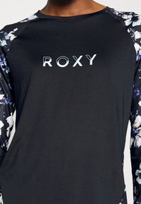Mustapaita pitkillä hihoilla, jossa on kukkakuosiset raglan-hihat ja vaaleansininen "ROXY"-logo edessä. Pehmeää ja joustavaa kangasta.