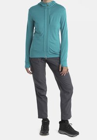 Giacca lunga turchese a maniche lunghe con colletto alto, zipper frontale e tasche laterali; abbinata a pantaloni grigi e scarponi da escursionismo neri.