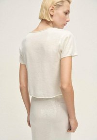 Maglione bianco corto a maglia con maniche corte e scollatura rotonda; presenta una texture liscia e una leggera lucentezza.