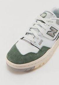Zapatilla blanca con detalles en ante verde, lengüeta de malla y suela de goma gris. Presenta un logotipo plateado y costuras decorativas. Número de modelo 550.