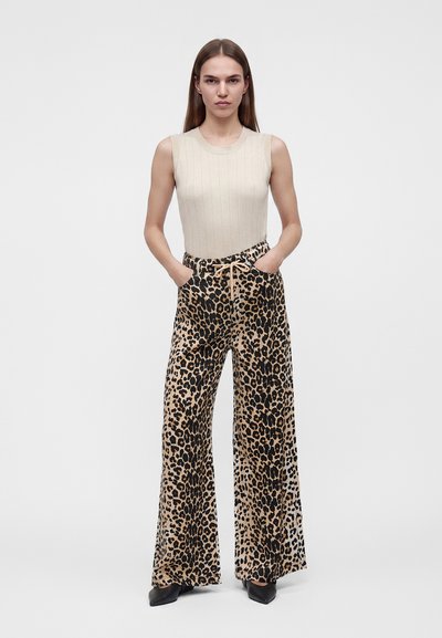 Femme portant un haut beige sans manches et un pantalon large imprimé léopard, les mains dans les poches, debout devant un fond clair uni.
