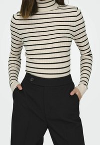 Pull à col roulé côtelé rayé crème et noir à manches longues, associé à un pantalon noir taille haute avec un détail bouton.