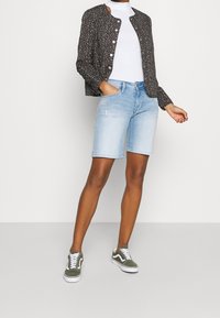 Svart jacka med blommönster och silverknappar, vit polotröja, ljusblå denim-shorts och gröna mockaskor med vita sulor.