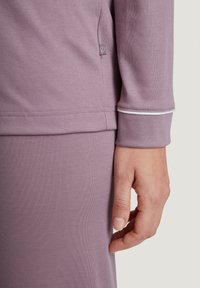 Long-sleeve lavender top, soft fabric with a slight texture. Features a white accent at the cuff and a small logo tag on the side.