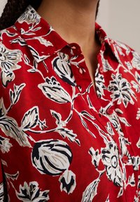 Röd blommig blus med krage, knappstängning och mönster av vita och svarta blommor. Tillverkad av lättviktigt tyg.