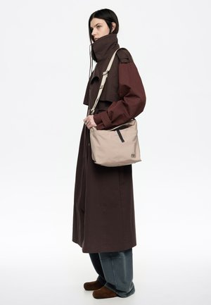 Bolso bandolera beige con un acabado suave y texturizado. Cuenta con cierre superior de cremallera, dos bolsillos laterales y una correa ajustable. La etiqueta muestra el nombre de la marca.