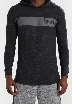 Mężczyzna noszący czarną, sportową koszulkę z długim rękawem i kapturem, z szarym poziomym paskiem i logo Under Armour na klatce piersiowej.