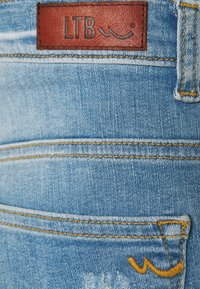Modré denimové džíny mají v pase hnědou koženou nášivku s nápisem "LTB", žluté švy a mírně opotřebovaný vzhled.