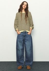 Pruhovaný nadrozmerný sveter v krémovej a hnedej farbe, skombinovaný so širokými modrými džínsami a hnedými mokasínami. Jednoduchý dizajn a voľný strih.