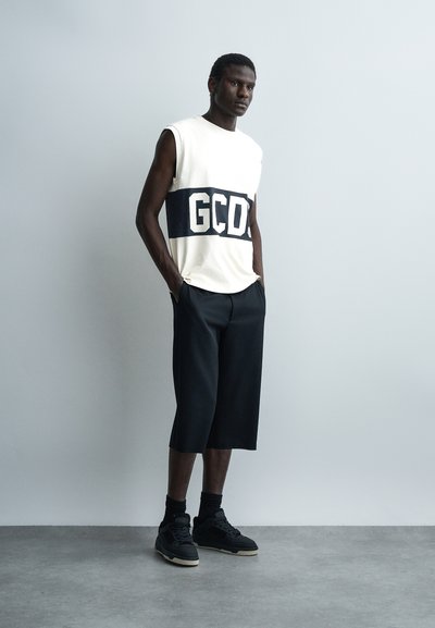 Άνδρας που φοράει λευκή αμάνικη μπλούζα με μαύρο λογότυπο "GCDS", μαύρο φαρδύ παντελόνι cropped, μαύρες κάλτσες και μαύρα αθλητικά παπούτσια, στέκεται με τα χέρια στις τσέπες.