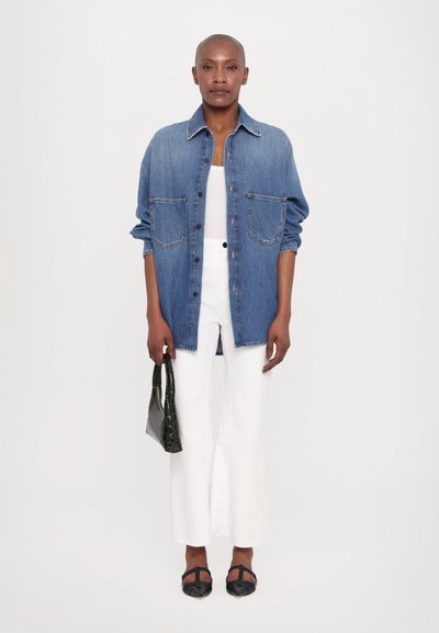 Denim shirt in medium blue with open front, two chest pockets, and rolled sleeves, paired with white wide-leg pants and a black handbag.