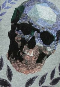 Grafische T-shirt met een geometrisch schedelontwerp, versierd met meerkleurige rhinestones, tegen een lichtgrijze achtergrond met donkere bladeren.