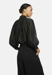 Black sheer blouse with gathered back, accented by white polka dots, oversized sleeves, and buttoned cuffs, paired with black tailored pants.