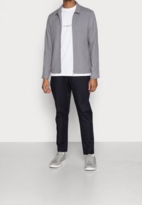 Ljusgrå zip-up jacka över en vit t-shirt med en logotyp, kombinerad med mörka byxor och grå sneakers. Enkel design, avslappnad passform.