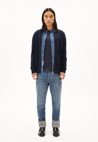 Marine cardigan over een denim shirt, gecombineerd met donkere denim jeans. De outfit heeft een casual pasvorm, met opgerolde mouwen en zwarte schoenen.