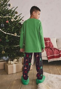 Long-sleeve green shirt, patterned pajama pants with festive graphics, and fuzzy green slippers. Set against a decorated Christmas tree.
