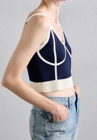 Top corto azul marino con ribete marfil, que presenta un escote en V, tirantes finos y tejido texturizado. El diseño incluye patrones circulares y curvados.