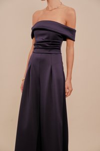 Fioletowa sukienka z odkrytymi ramionami, dopasowaną górą, plisowanymi szerokimi spodniami oraz gładką fakturą materiału. Minimalistyczny design bez wzorów.