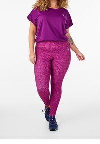 Fioletowa koszulka sportowa z krótkim rękawem, zestawiona z dopasowanymi różowymi legginsami w wzór zebry. Noszona z niebieskimi sneakersami.