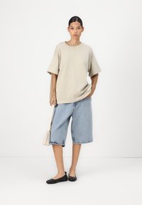 Beige oversized t-shirt, denim shorts die boven de knie eindigen, zwarte platte schoenen en een kleine lichte tas. Geweven visnet mouwen zichtbaar.