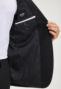 Schwarzer Blazer mit glatter Außenseite und gemustertem Futter. Verfügt über ein Taschenlabel und ein maßgeschneidertes, schlank geschnittenes Design.