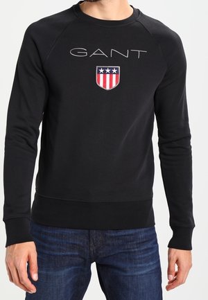 Czarna bluza z napisem "GANT" i czerwoną, białą i niebieską tarczą z gwiazdkami, noszona przez osobę w niebieskich dżinsach.