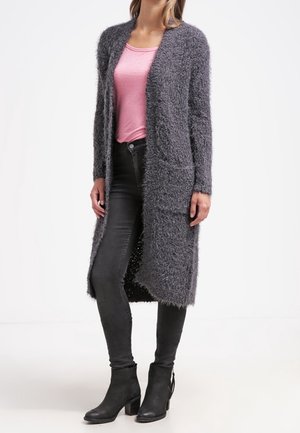 Kvinde iført en lang grå fnugget cardigan over en lyserød skjorte, sorte smalle jeans og sorte ankelstøvler, stående mod en hvid baggrund.