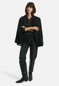 Schwarzer maßgeschneiderter Mantel über einem braunen strukturierten Pullover, kombiniert mit schwarzen engen Jeans und kniehohen, glänzenden schwarzen Stiefeln mit Reißverschlüssen.