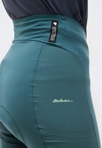 Primer plano de una persona que lleva leggings deportivos de talle alto en color verde azulado, con un pequeño logo en la cadera y una etiqueta negra en la cintura.