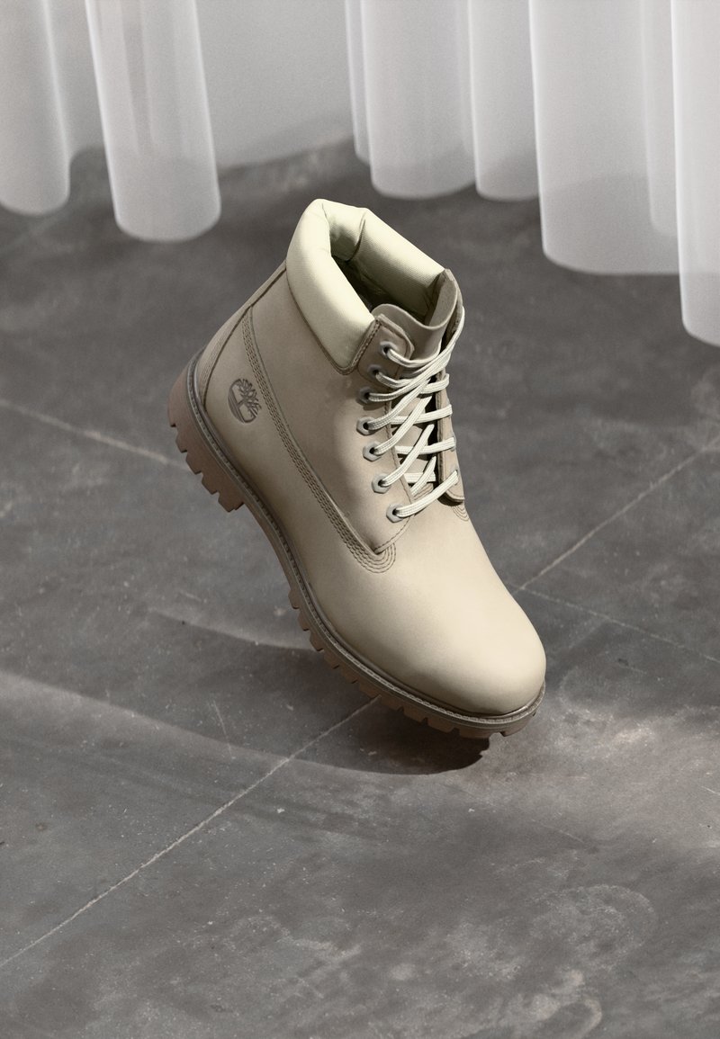 Light beige leather ankle boot with padded collar, laces, and rugged sole on a gray tiled floor beneath sheer white curtains.