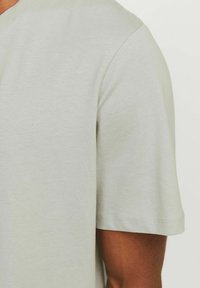 Hellbeige T-Shirt mit kurzen Ärmeln, getragen von einer Person mit dunkler Haut, von der Schulter bis zur Mitte des Oberkörpers vor einem schlichten Hintergrund gezeigt.