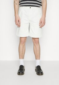 Hvide shorts med lige snit, lavet af bomuldsstof, med sidelommer og en enkelt knaplukning, parret med sorte sko og hvide sokker.