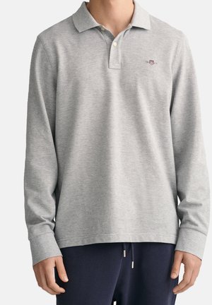 Personne portant un polo à manches longues gris clair avec un petit emblème sur la poitrine et un pantalon bleu marine avec des cordons.