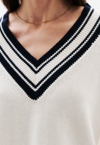 Maglione a V in crema con costine a righe blu navy e bianche. Presenta un dettagliato lavorato a maglia e cuciture visibili.
