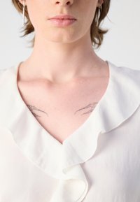 Hvid bluse med en volangkrave, glat tekstur og V-halsdesign. Synlige tatoveringer på brystområdet under kraven.
