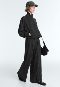 Femme portant un pantalon large à carreaux noirs et une veste assortie à col montant, une casquette noire, des chaussures noires et un sac à épaule noir.