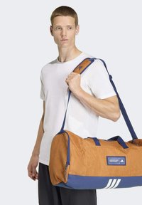 Borsa duffle in tessuto arancione con base e tracolla blu. Presenta tre strisce bianche e un'etichetta con logo sul lato. Texture liscia e forma strutturata.