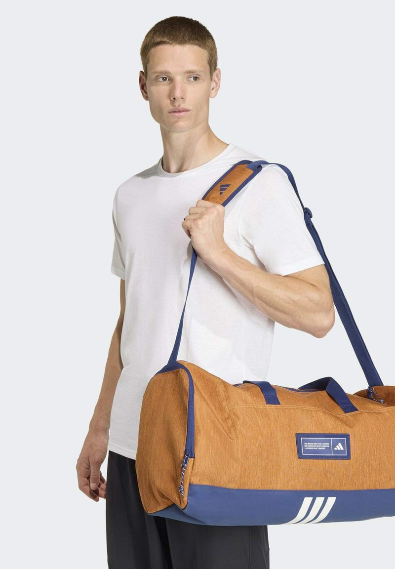 Borsa duffle in tessuto arancione con base e tracolla blu. Presenta tre strisce bianche e un'etichetta con logo sul lato. Texture liscia e forma strutturata.