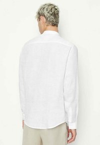Camicia bianca in lino a maniche lunghe con vestibilità rilassata, orlo rotondo e polsini con bottone. Presenta una texture liscia e un design minimalista.