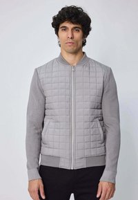 Veste bombardier grise avec un devant matelassé, des manches côtelées et une fermeture éclair sur le devant. Elle présente une finition texturée et des poches latérales.