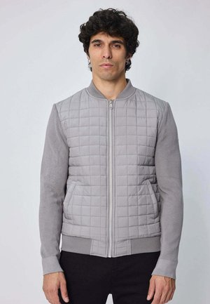 Veste bombardier grise avec un devant matelassé, des manches côtelées et une fermeture éclair sur le devant. Elle présente une finition texturée et des poches latérales.