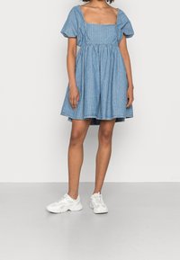 Mujer con un vestido corto azul de mangas abullonadas y escote cuadrado, llevando zapatillas blancas de suela gruesa, de pie sobre un suelo gris.