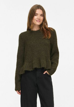 Femme portant un pull duveteux vert foncé à manches longues et un pantalon noir taille haute, debout, une main dans la poche.