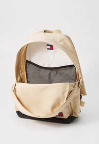 Rucsac bej cu o suprafață moale și texturată, având un buzunar din plasă în interior și o secțiune inferioară neagră. Logo-ul este vizibil pe exterior.