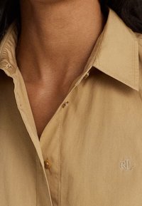 Beige košile na knoflíky s špičatým límcem, která má jedno zapínání na knoflík a vyšitým logem 'RL' na hrudi. Bavlněná tkanina.