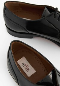 Crne kožne cipele s sjajnim zavr�šetkom, dizajn vezanja i smeštem od smeđe kože. Oznaka "Next Signature Italian Leather" vidljiva unutra.