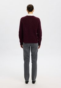 Bordo spalvos megztinis su ilgomis rankovėmis, apvalia apykakle ir laisvo silueto, derinamas su pilkomis aukšto liemens džinsinėmis kelnėmis, matomas iš nugaros.