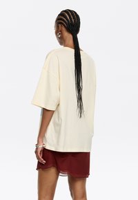 Kremowy oversize'owy t-shirt z krótkimi rękawami, charakteryzujący się luźnym krojem. Połączony z bordową, przezroczystą spódnicą, podkreślającą warstwowe faktury.
